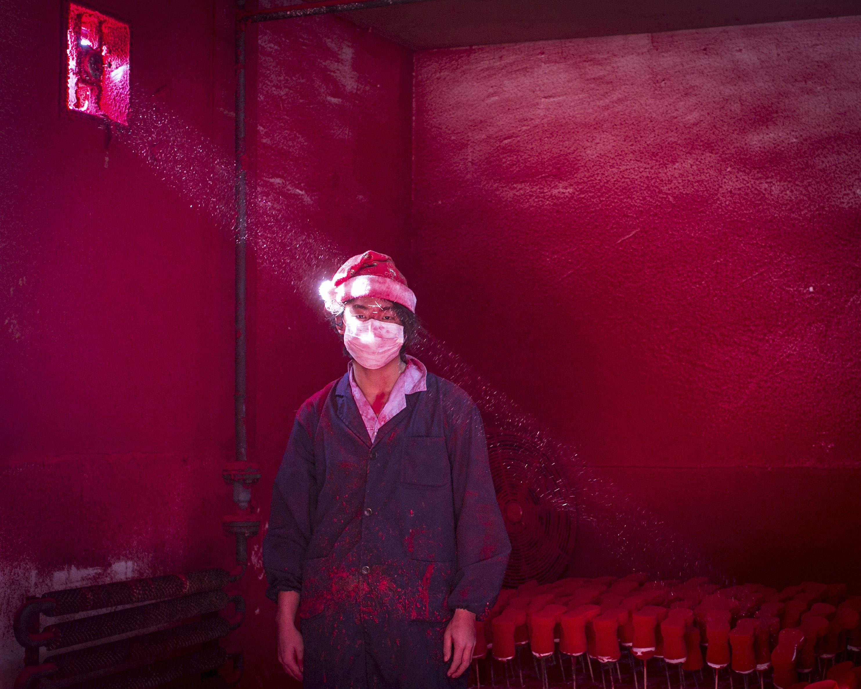 Rongui Chen | Φωτογραφία 19χρονου Κινέζου εργάτη με μάσκα στο πρόσωπο και σκούφο του Άγιου Βασίλη ενώ στέκεται δίπλα σε χριστουγεννιάτικα στολίδια που στεγνώνουν σε εργοστάσιο και κόκκινη σκόνη που χρησιμοποιήθηκε για τον χρωματισμό αιωρείται στον αέρα