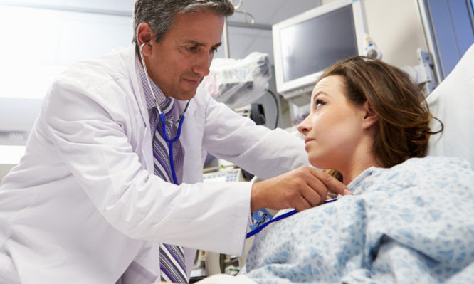 Male Doctor Examining Laid Down Female Patient In Emergency Room