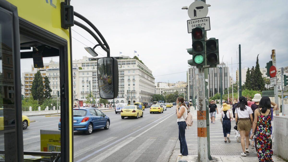 Woman waiting at pedestrian crossing talking on mobile phone while vehicles and a bus pass in July 2019 Athens Greece.