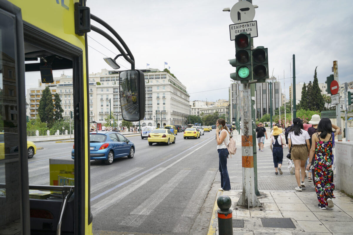 Woman waiting at pedestrian crossing talking on mobile phone while vehicles and a bus pass in July 2019 Athens Greece.