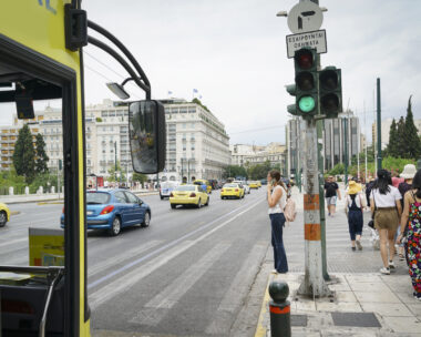 Woman waiting at pedestrian crossing talking on mobile phone while vehicles and a bus pass in July 2019 Athens Greece.
