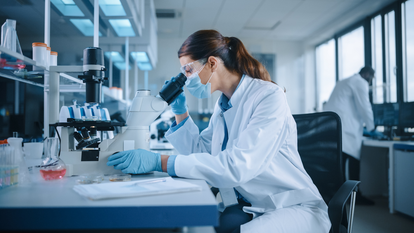 Female Scientist in Face Mask and Glasses Looking a Petri Dish with Genetically Modified Sample Chemicals Under a Microscope. Microbiologist Working in Modern Laboratory with Technological Equipment.