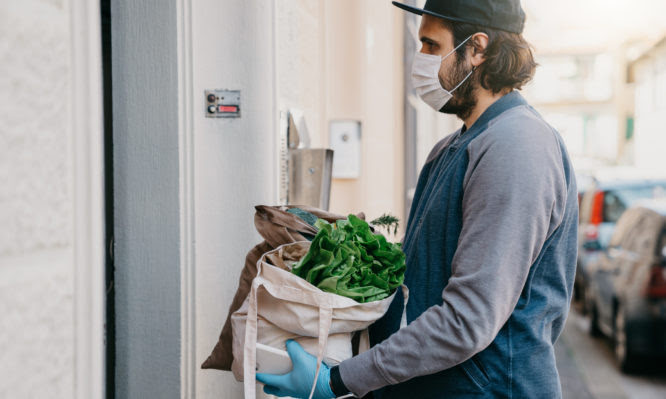 A man is delivering a bag of vegetables and fruit.  He's waiting for the customer to open the door.
