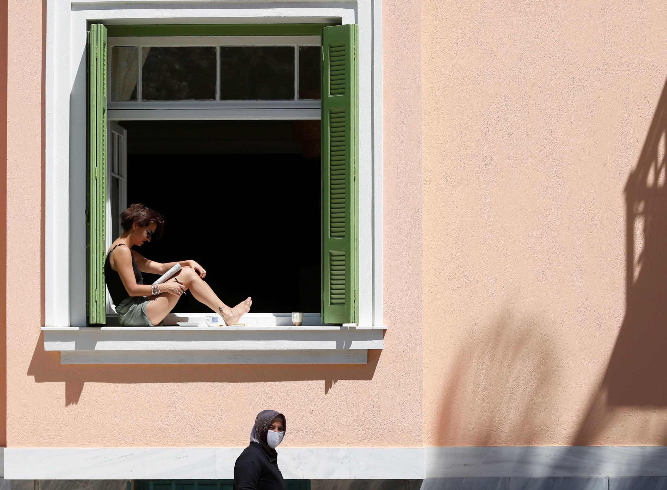 A woman reads as she sits in a window as another woman carries bags after shopping at an open market following the coronavirus disease outbreak (COVID-19) ahead of Greek Orthodox Easter in Athens, Greece, April 17, 2020. REUTERS
