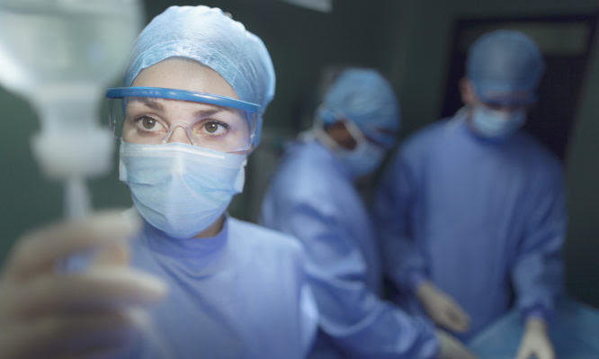 Female woman nurse OR staff surgeon touching adjusting checking intravenous bottle fluid dressed in safety gown scrubs and performing operation in operating room medical facility hospital healthcare determination preparation