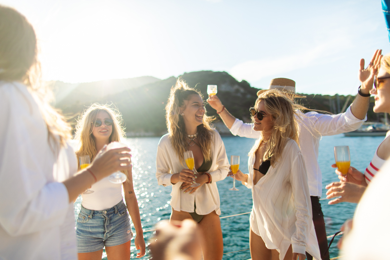 Group of beautiful young people having a party on sailboat deck before sunset. They are dancing, drinking wine and cocktails, relaxing and enjoying their summer vacation on sailing yacht