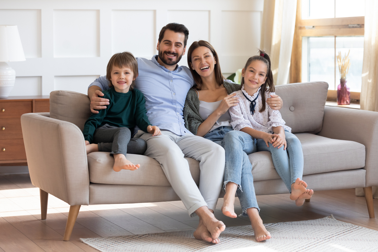 Portrait of happy young Caucasian family renters with two little children relax on couch in new modern home. Smiling parents with small kids sit rest on sofa in renovated design house. Rent concept.