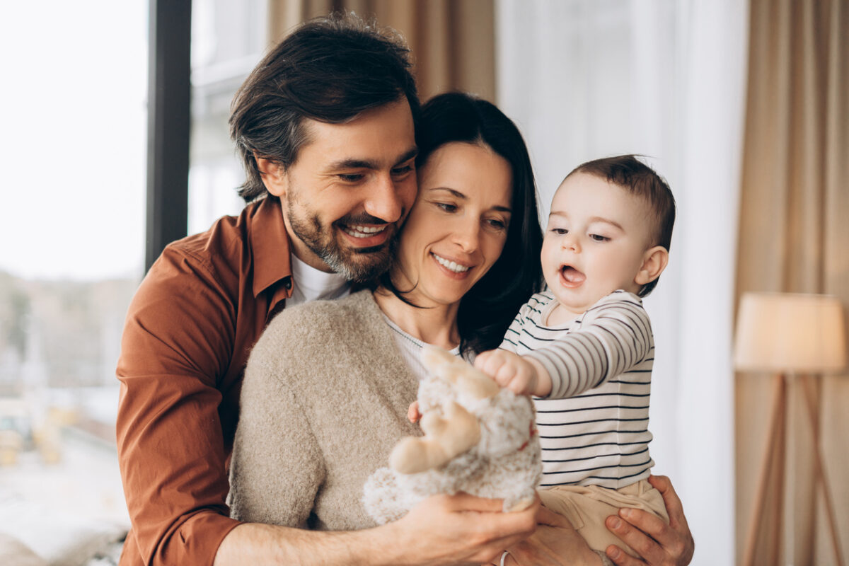 Smiling parents holding their baby son, who is reaching for a cuddly teddy bear, are enjoying precious moments together in the cozy comfort of their home, radiating love and happiness. Family concept