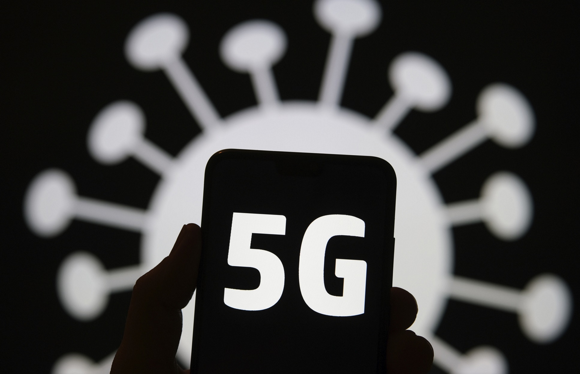 5G letters on smartphone silhouette hold in a hand and blurred coronavirus image on the blurred background. Real photo, not a a montage. (5G letters on smartphone silhouette hold in a hand and blurred coronavirus image on the blurred background. Real