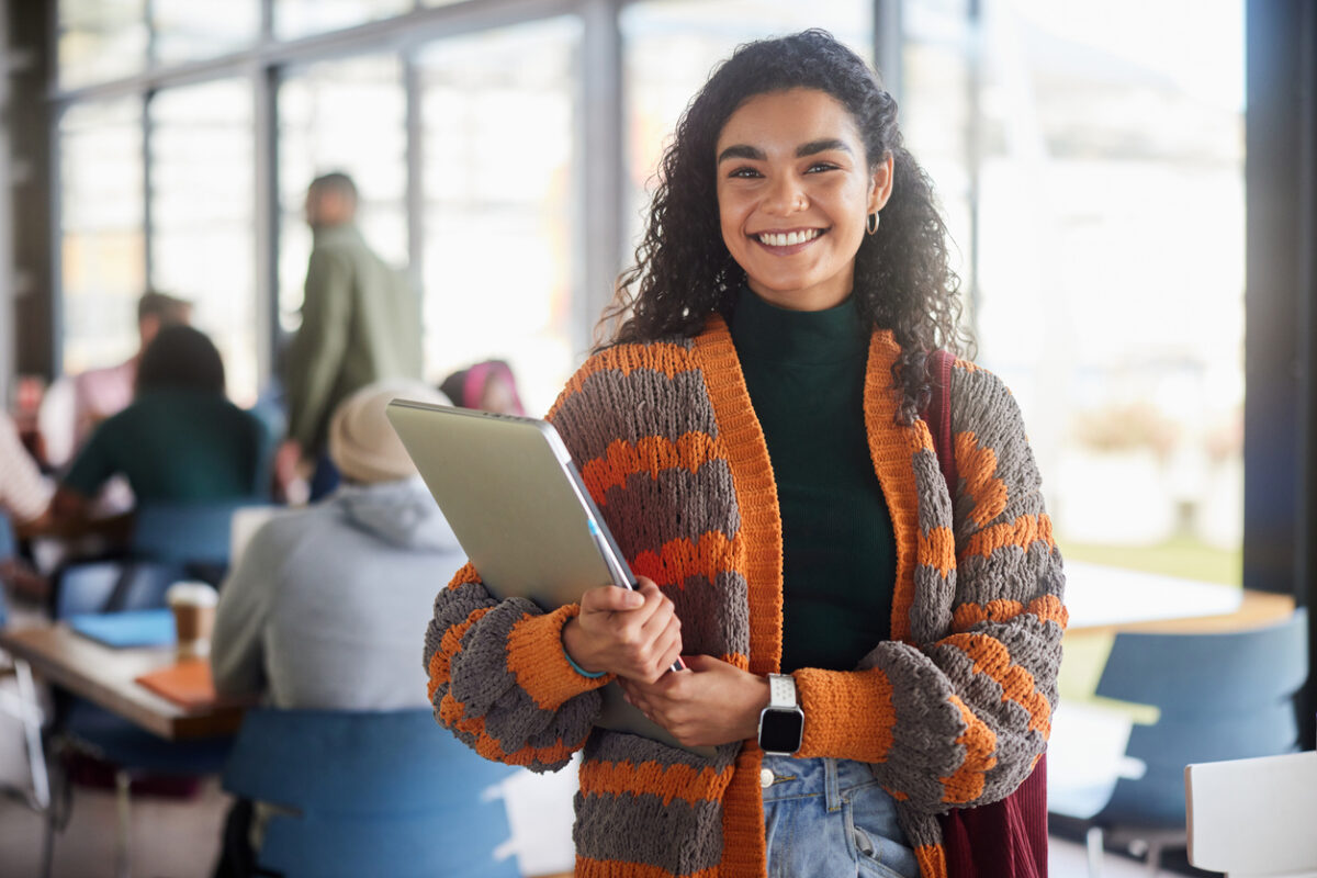 Portrait of a young female college student smiling while standing with her laptop in a cafeteria at school