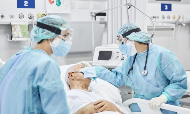 Female doctor talking with patient along coworker in ICU. Man is lying on bed amidst essential workers. Healthcare workers are in protective workwear.