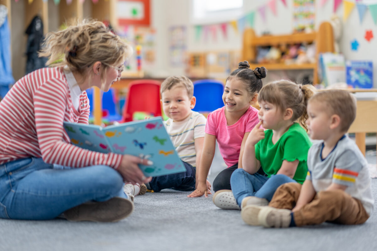 A preschool teacher sits on the floor of her classroom with a small group of students as she reads them a book. The children are each dressed casually and are focused on the story.