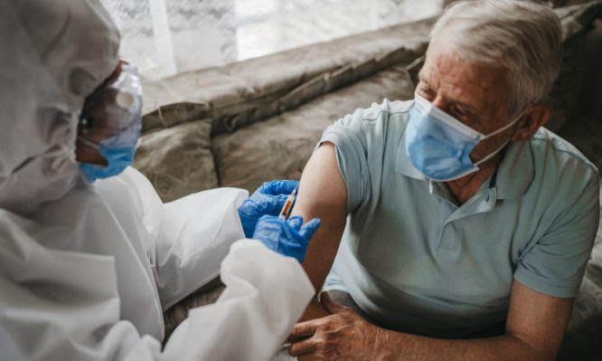 Female doctor in protective suit giving vaccine against covid19 virus or injection to senior man at home. Therapy at home in pandemic conditions