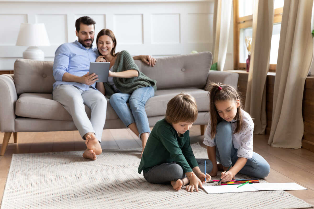 Cute little brother and sister sit on floor drawing in album, young parents relax on comfortable couch in living room using tablet, young Caucasian family rest together enjoy leisure weekend at home