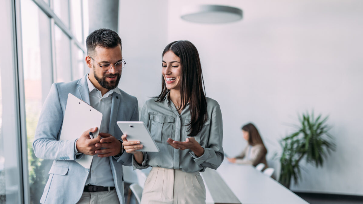 Shot of a two confident business persons talking in the work place. Two colleagues using a digital tablet while walking in a modern office. Businessman and businesswoman in meeting discussing business strategy. Business coworkers working together in the offic