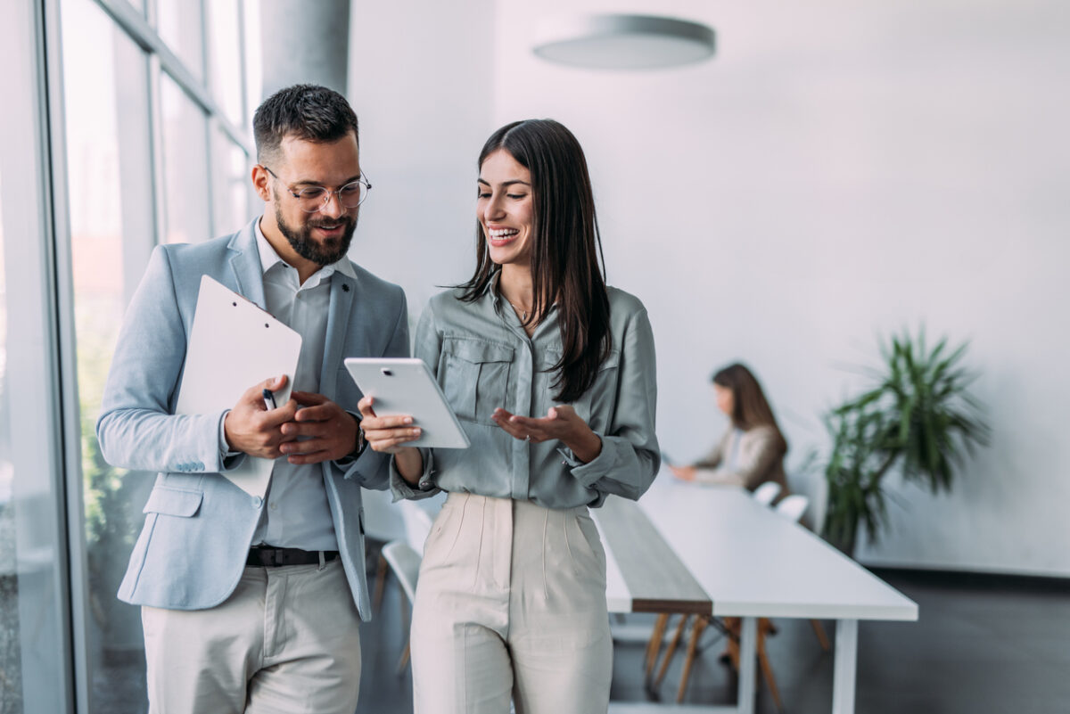 Shot of a two confident business persons talking in the work place. Two colleagues using a digital tablet while walking in a modern office. Businessman and businesswoman in meeting discussing business strategy. Business coworkers working together in the offic