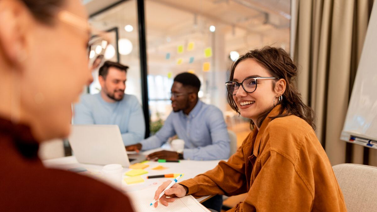 Young businesswoman taking notes and smiling during a collaborative meeting with colleagues in a modern office, embodying teamwork and fostering innovative ideas for project success