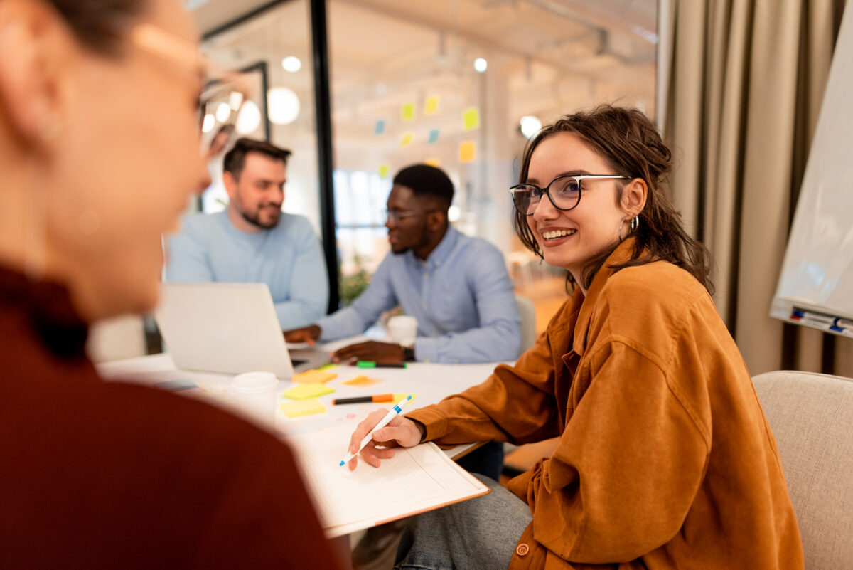 Young businesswoman taking notes and smiling during a collaborative meeting with colleagues in a modern office, embodying teamwork and fostering innovative ideas for project success