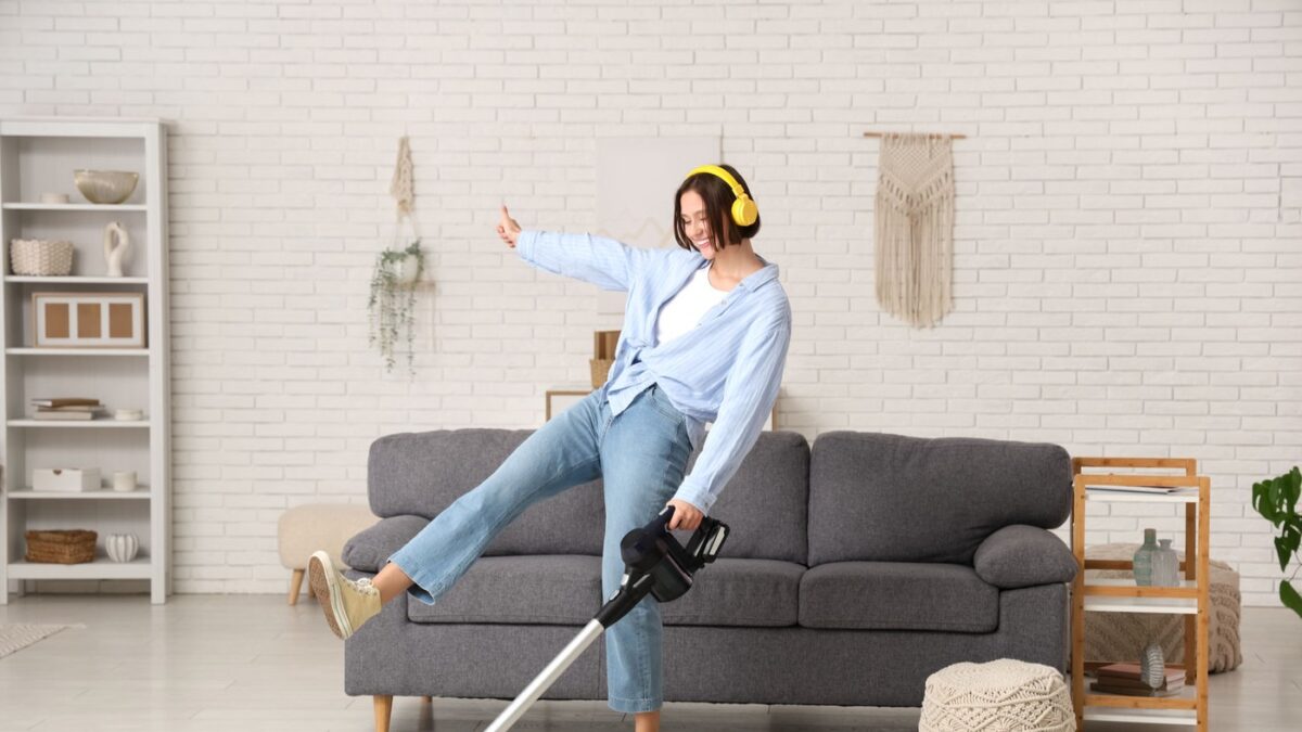 Beautiful young woman in headphones having fun with modern vacuum cleaner at home