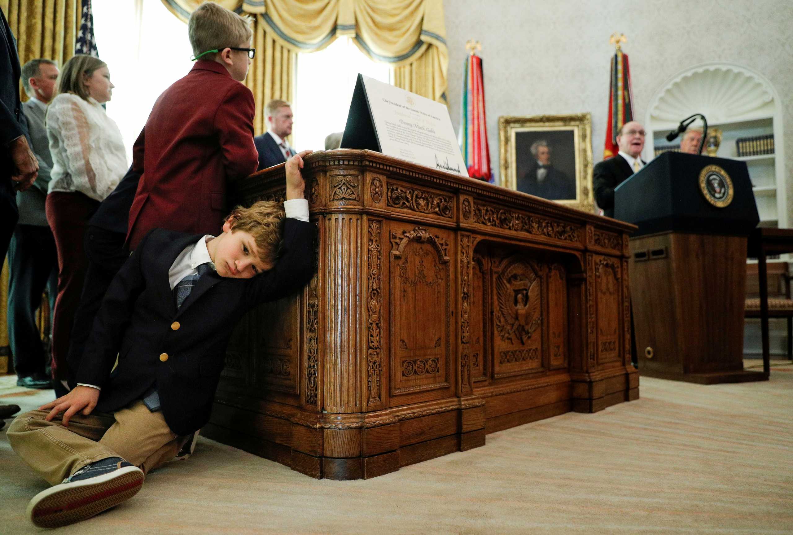 Sammy Olszta, 6, grandson of U.S. Olympic wrestling Gold Medalist and Presidential Medal of Freedom recipient Dan Gable, leans against the Resolute Desk as Gable delivers remarks beside U.S. President Donald Trump during a presentation ceremony inside the Oval Office at the White House in Washington, U.S., December 7, 2020. REUTERS