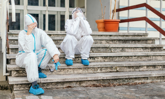 Two female health worker resting outdoors at hospital backyard and taking off her protective mask and suit. They are under big pressure and stress during corona virus pandemic