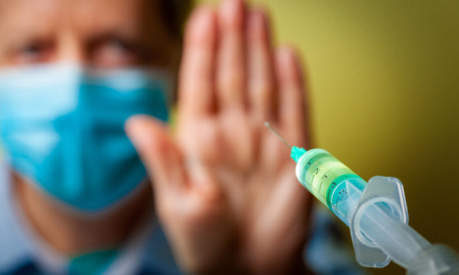 Vaccine-hesitant adult man wearing surgical protective face mask in the background holding up hand as a stop sign against a SARS-CoV-2 injection syringe approaching from the foreground