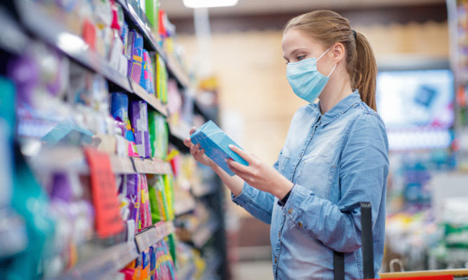 Young woman wearing protective face mask, looking at box of sanitary pads in supermarket aisle, COVID-19 pandemic era