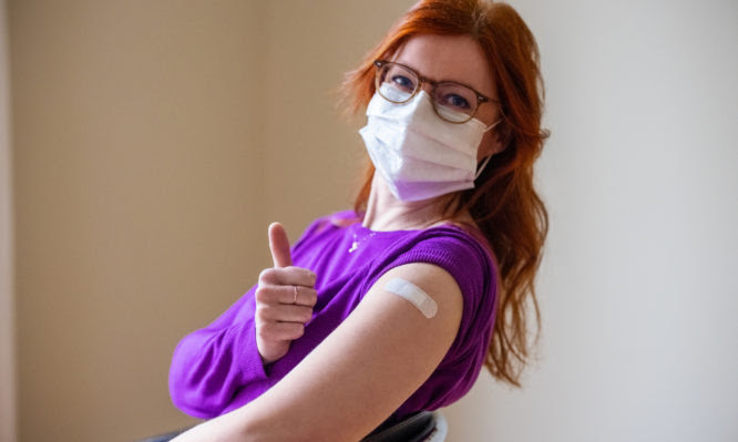 Woman wearing face mask looking at camera showing thumbs up after getting the covid-19 vaccine. Female gesturing thumbs-up sign after getting coronavirus vaccine at vaccination center.