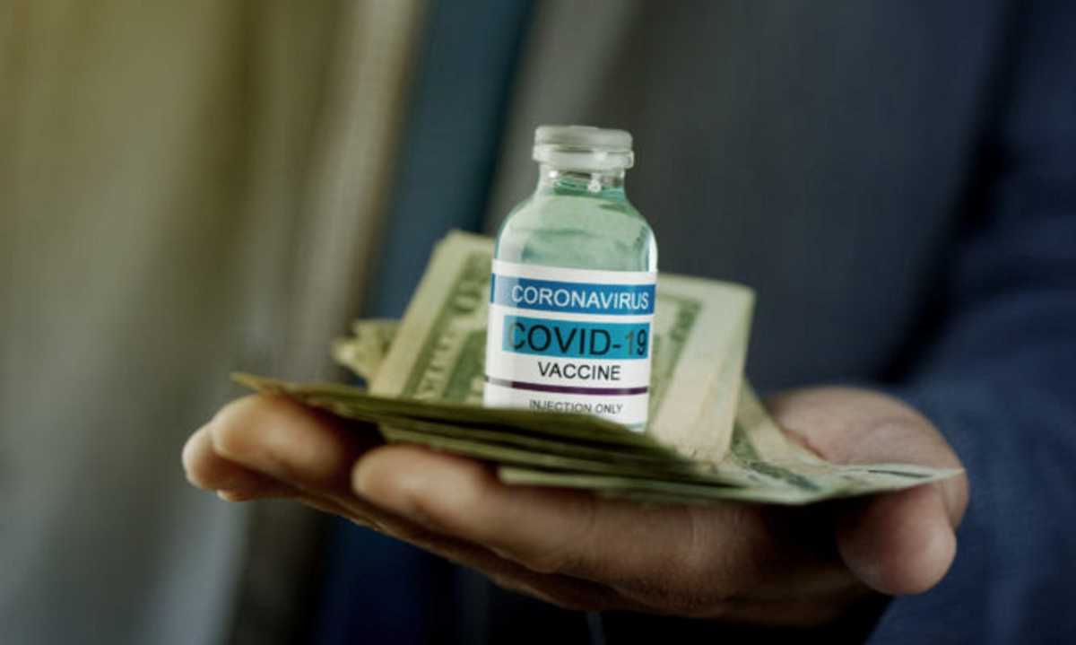 a businessman, wearing a grayish-blue suit jacket and a blue necktie, has a simulated covid-19 vaccine bottle and a bunch of dollar notes in his hands