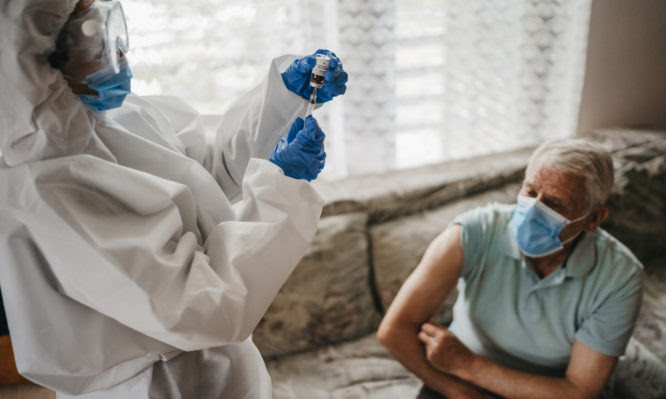 Female doctor in protective suit giving vaccine against covid19 virus to senior man at home