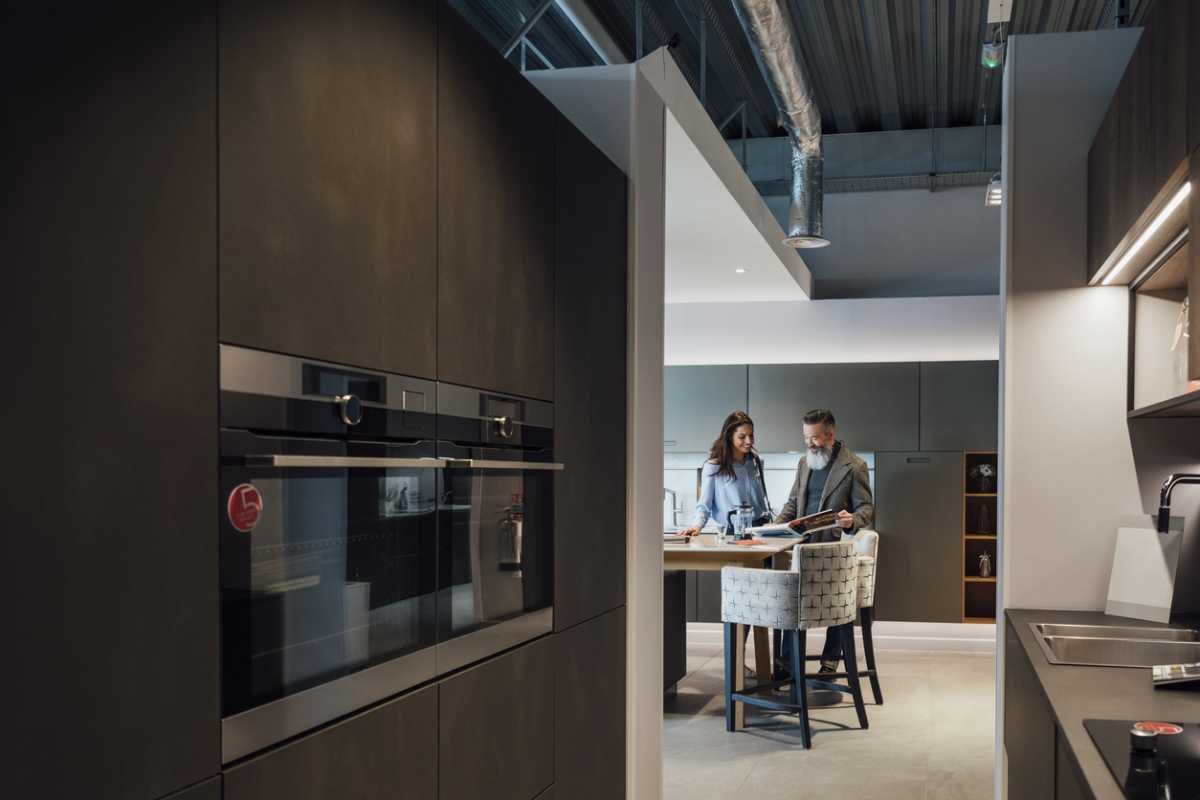 A wide-view shot of a mid-adult woman viewing a modern kitchen showroom interior with her father, she is considering buying a new kitchen to improve her home. They are reading through an in-store leaflet.