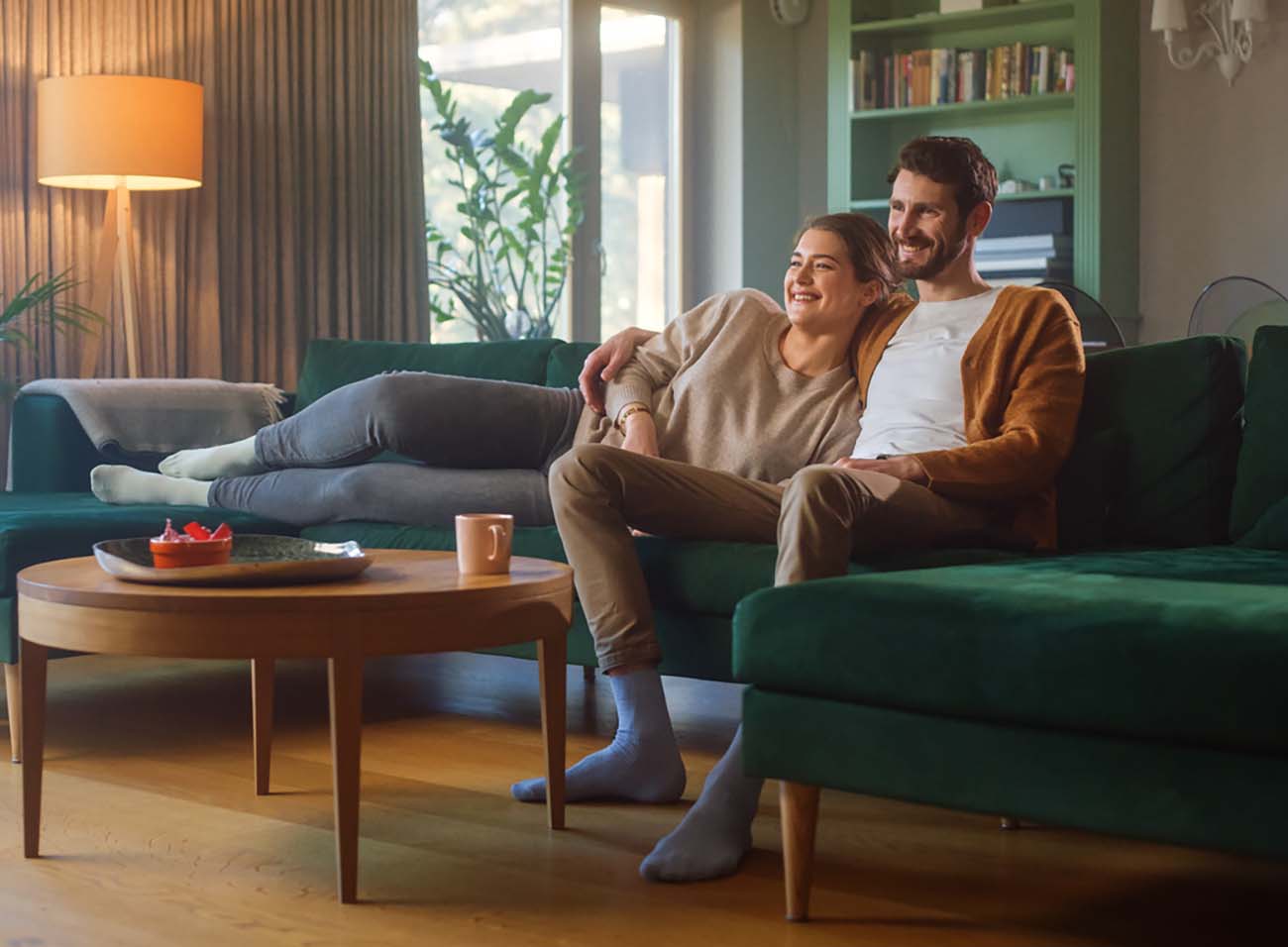 Couple Watches TV together while Sitting on a Couch in the Living Room. Girlfriend and Boyfriend embrace, cuddle, talk, smile and watch Television Streaming Services. Home with Cozy Stylish Interior.