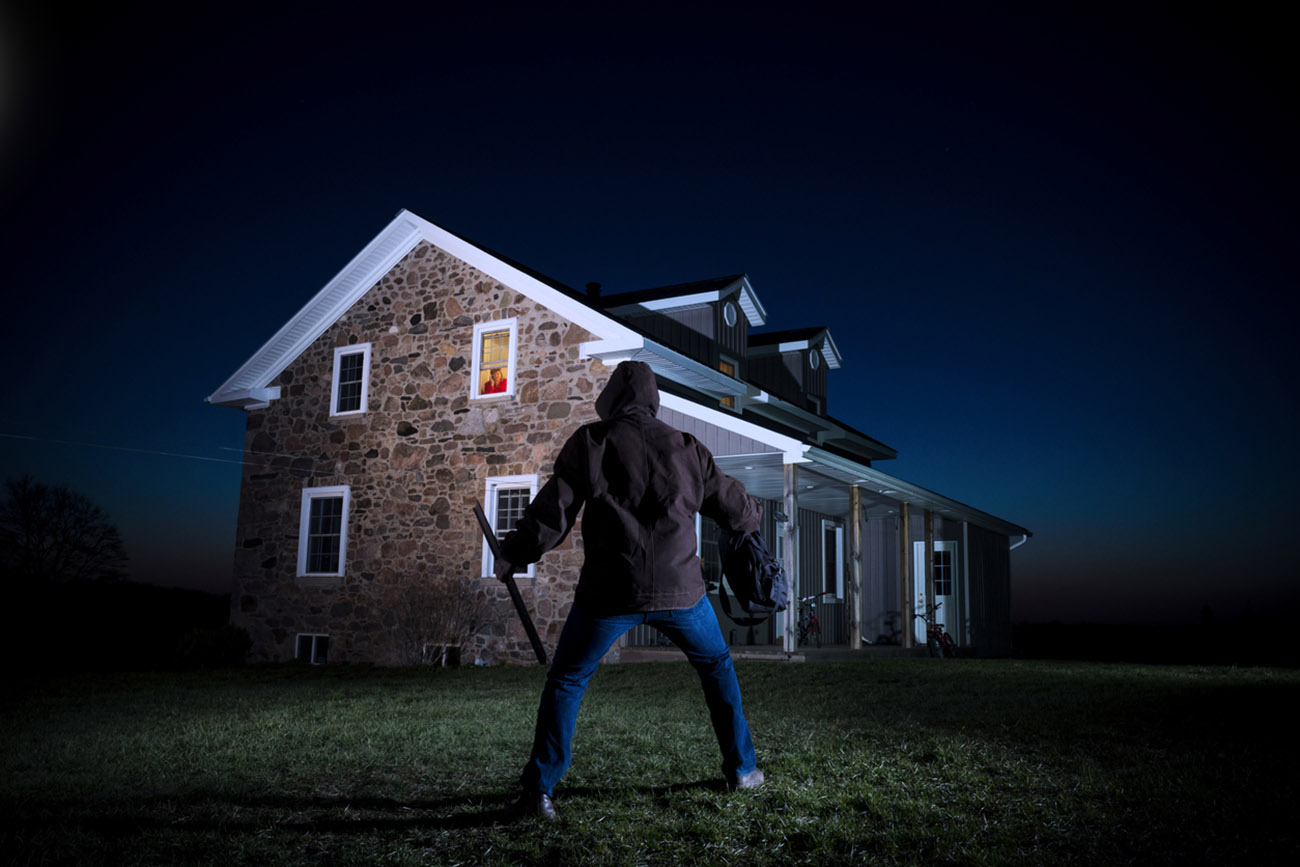 A night time photograph of a burglar outside house at night with a homeowner watching from a bedroom window while on the phone.Please browse: