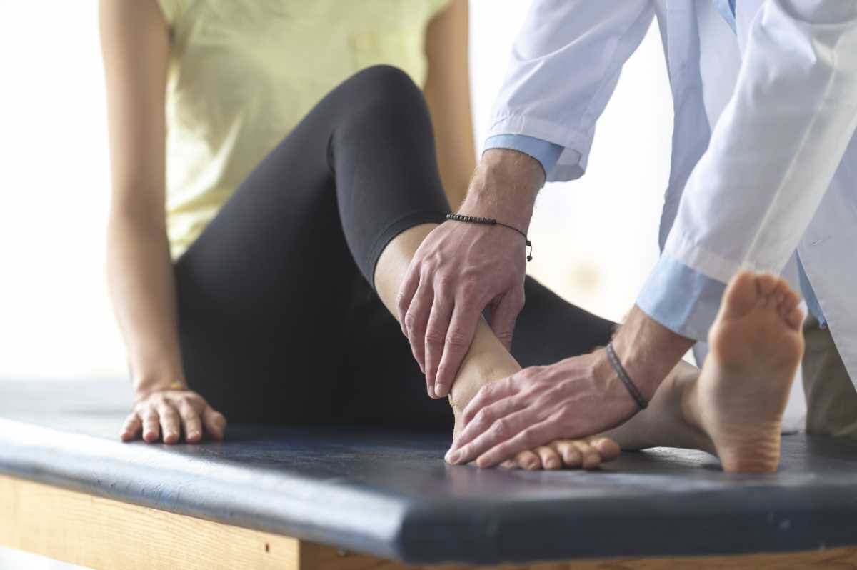 A woman is sitting on an examination table in a doctor's office. She has her shoes and socks off and the doctor is examining one of her ankles.