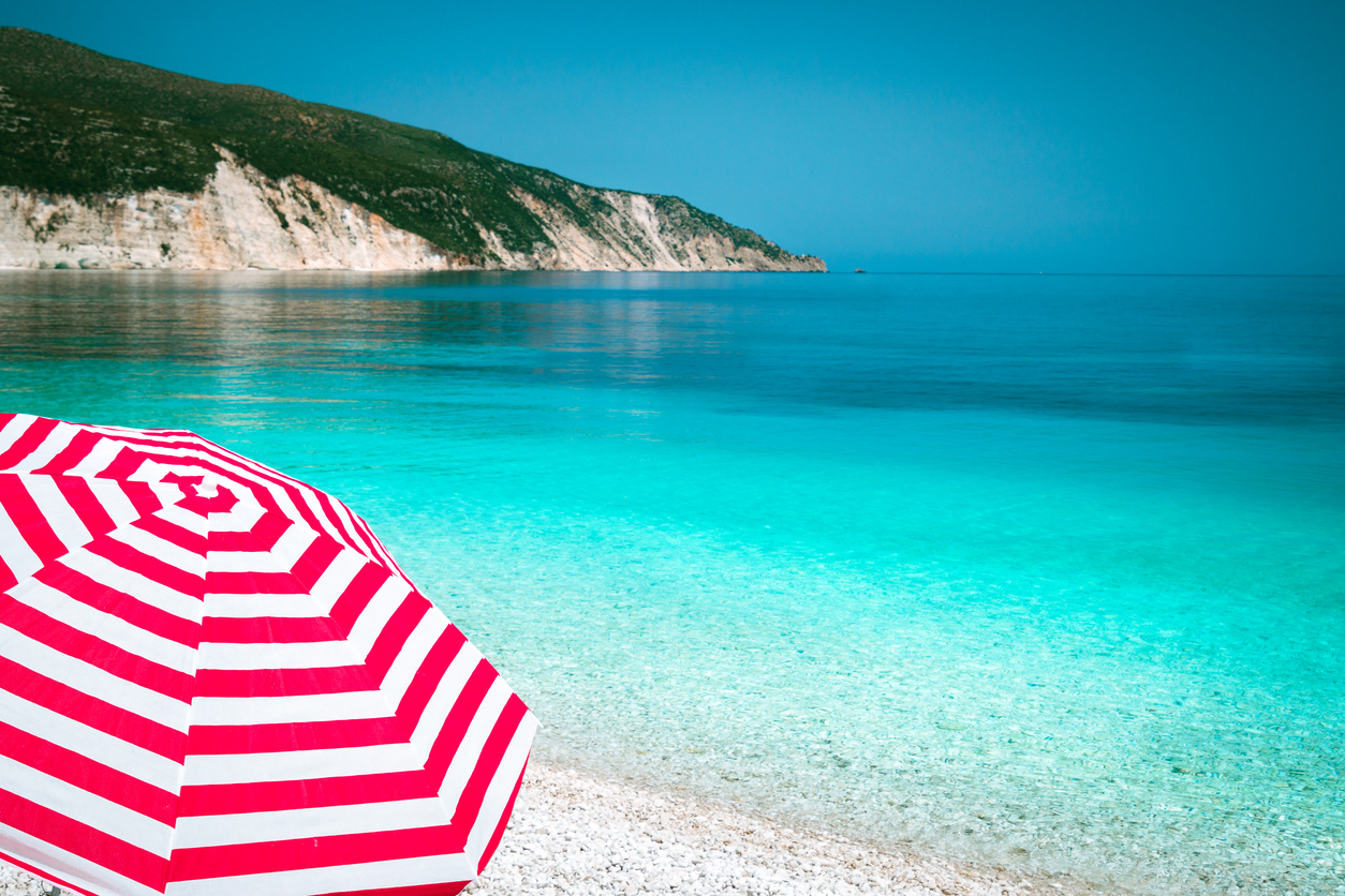 Striped red sun beach umbrella on a pebble beach with turquoise blue sea, white rocks and sky in background.