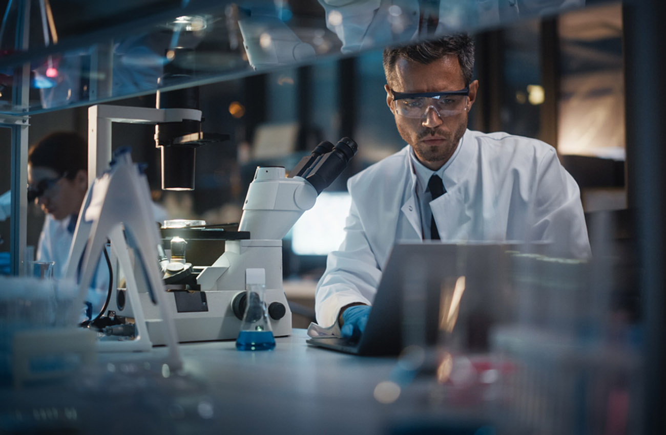Modern Medical Research Laboratory: Male and Female Scientists Work on Laptop, Do Data Analysis, Use Microscope. Advanced Pharmaceutical Lab for Medicine, Biotechnology Development. Evening Time