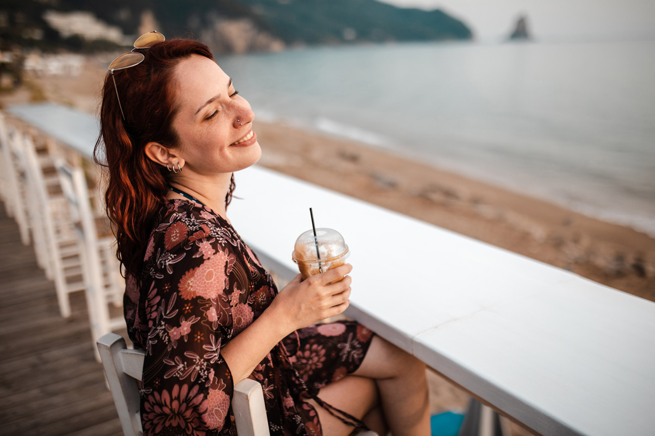 Cheerful young woman relaxing at the beach coffee shop, drinking the coffee and enjoying the view of the Ionian sea