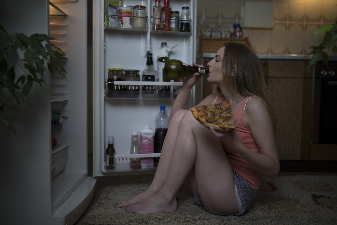 Young woman wearing pajamas sitting in front of the refrigerator, eating pizza slice and drinking wine late at night