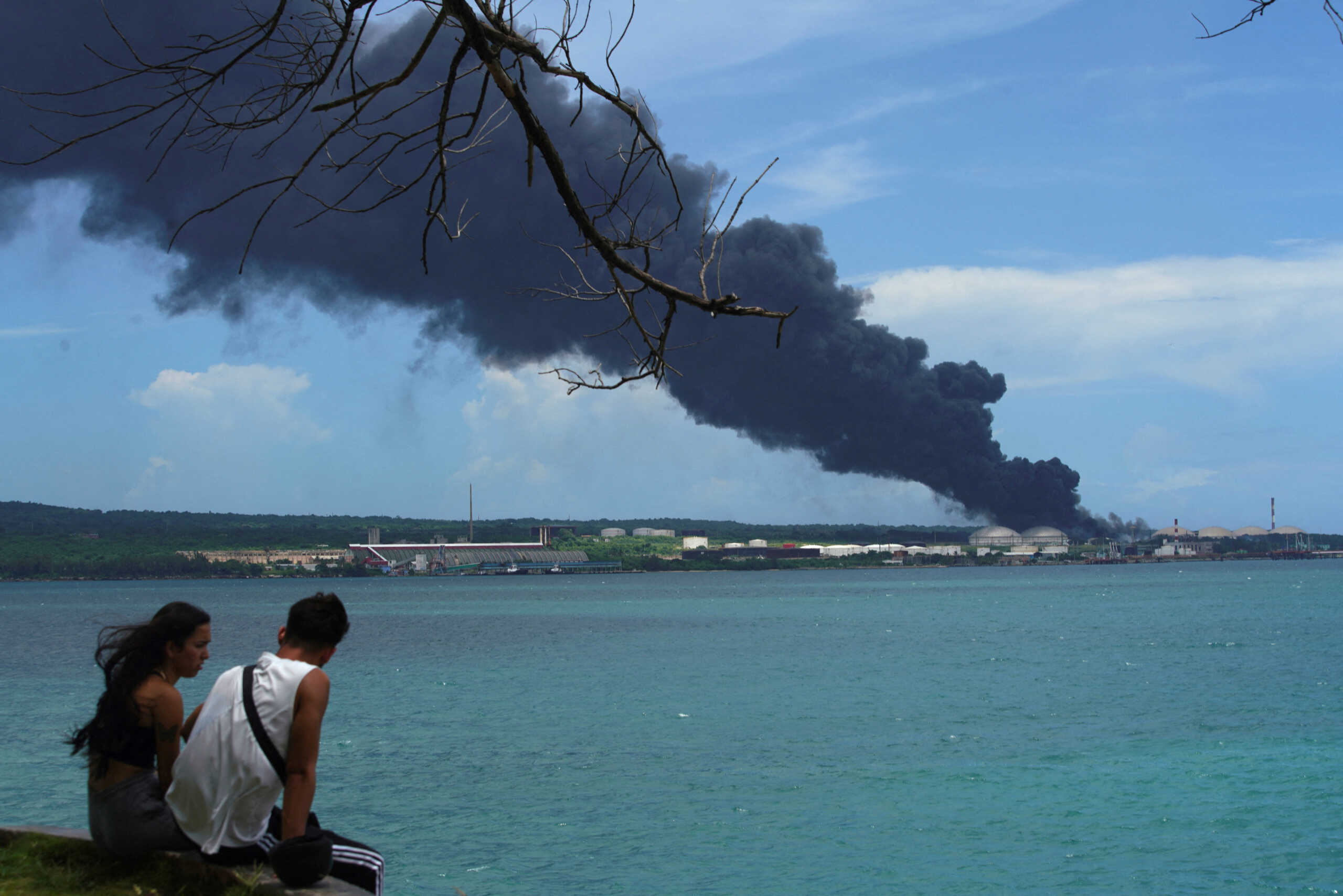 FILE PHOTO: A couple sits near the sea as smoke rises over fuel storage tanks that exploded near Cuba's supertanker port in Matanzas, Cuba, August 7, 2022. REUTERS
