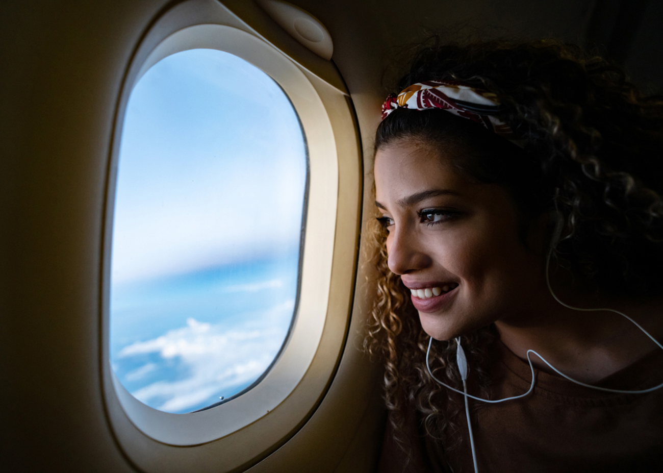 Portrait of a happy woman traveling by airplane and listening to music with headphones while looking through the window - lifestyle concepts