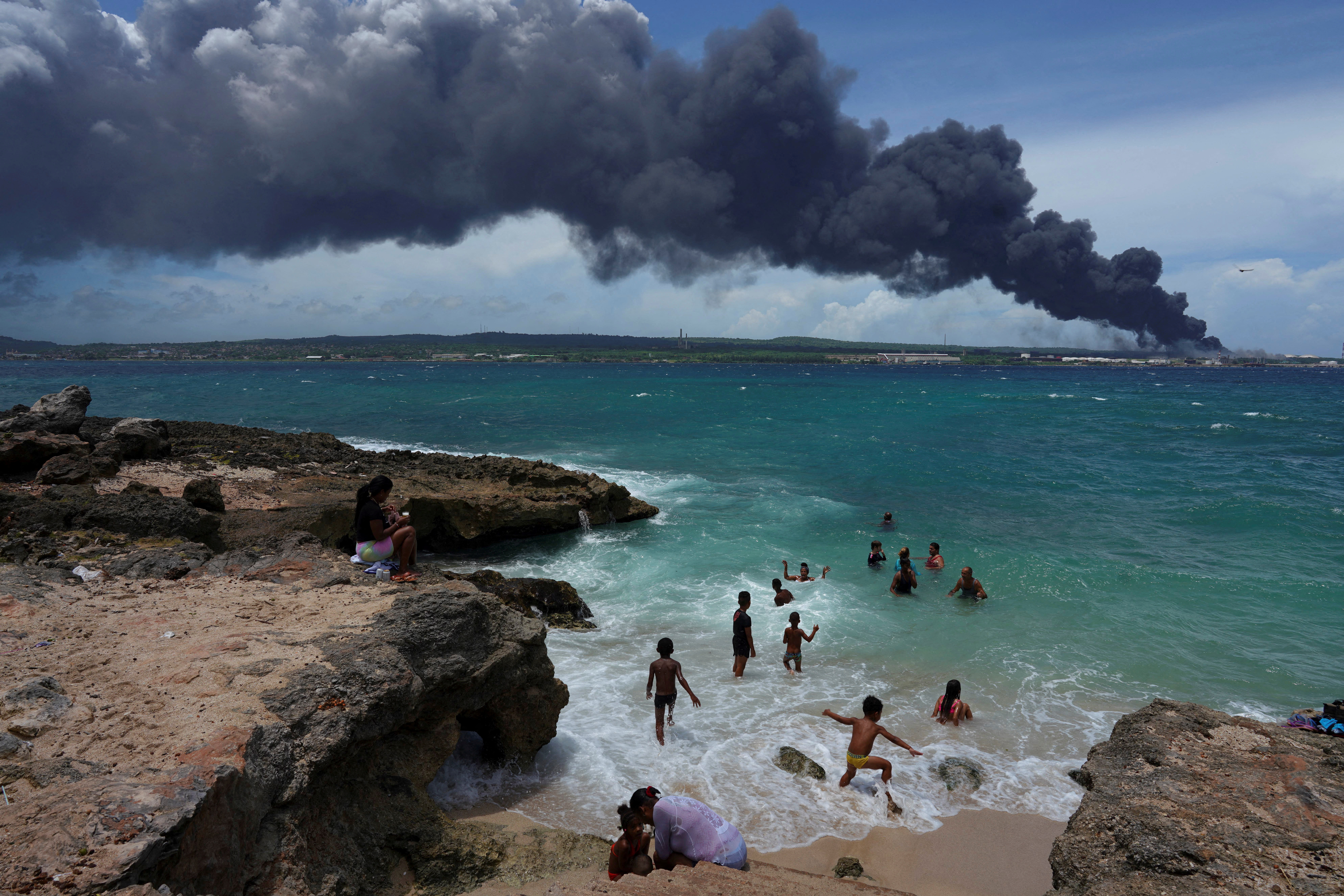 People enjoy the beach near the smoke of a fire over fuel storage tanks that exploded near Cuba's supertanker port in Matanzas, Cuba, August 6, 2022. REUTERS