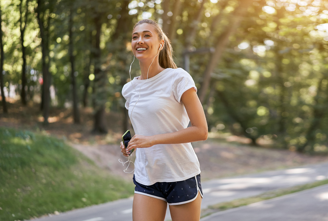Woman listen music running on asphalt track summer park Caucasian female earphones jogging sunny morning Runner training hold smartphone in hand Healthy lifestyle positive concept