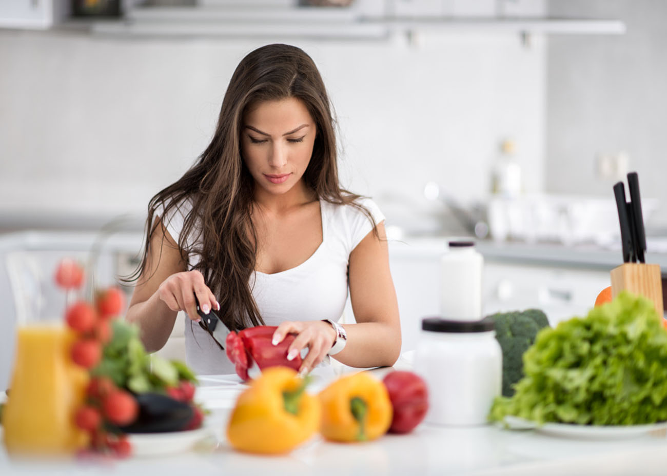 Brunette female cutting vegetables for salad that she is preparing in the kitchen. On the table are plate with green salad, broccoli, red and yellow bell peppers, tomatoes, some ingredients and orange juice. There are utensils and other kitchen elements in the background.