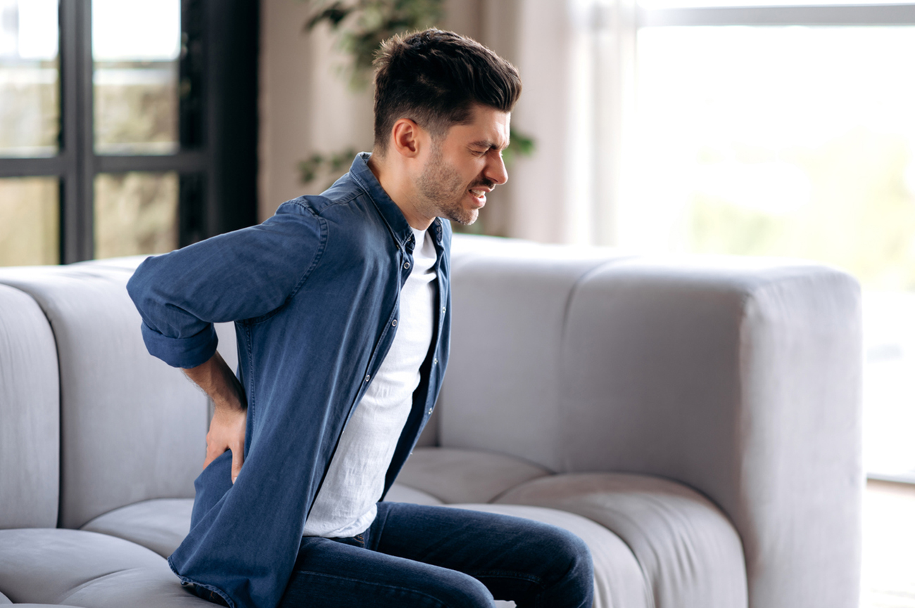 Sad upset young adult caucasian bearded man, in casual clothes, sitting on the sofa in the living room, massaging his back, suffering from back pain, suffering from arthritis, sciatica