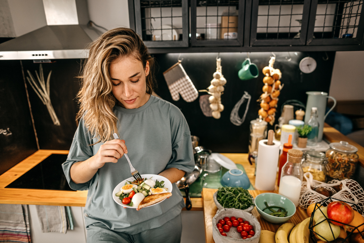 Young women sitting on stool in modern kitchen eating healthy breakfast of toast and eggs with salad using a fork at home