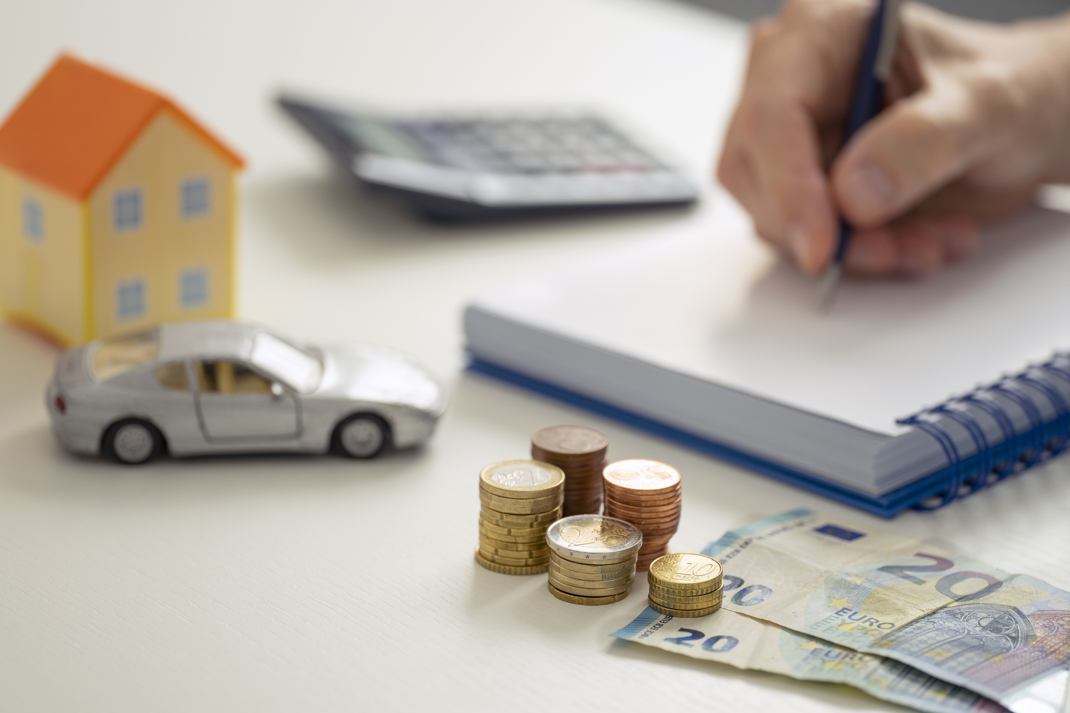 A MALE HAND WRITING IN THE NOTEBOOK NEXT TO A CALCULATOR, A LITTLE HOUSE, A TOY CAR AND SEVERAL STACKS OF EURO COINS AND NOTES. HOME ECONOMY CONTROL CONCEPT. FOCUS SELECTED.