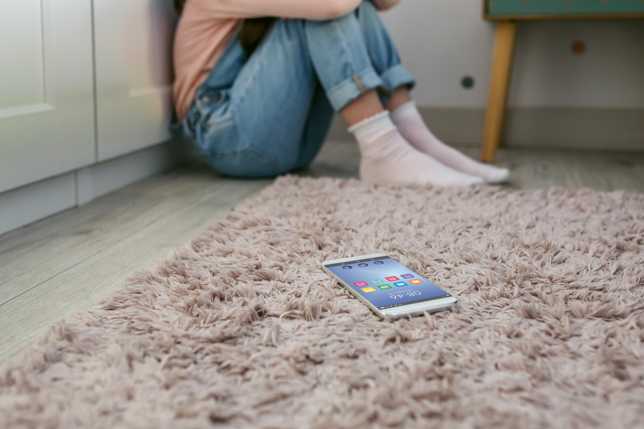 Mobile phone lying on the floor with unrecognizable sad little girl sitting behind. Selective focus on mobile in foreground