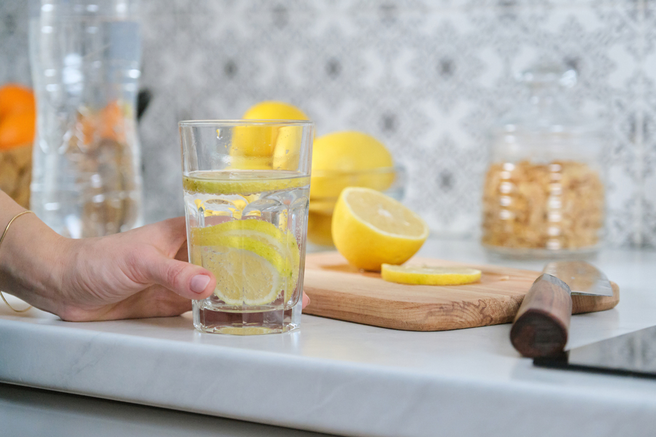 Female hand with slice of lemon in kitchen, with freshly made drink sparkling water with lemon