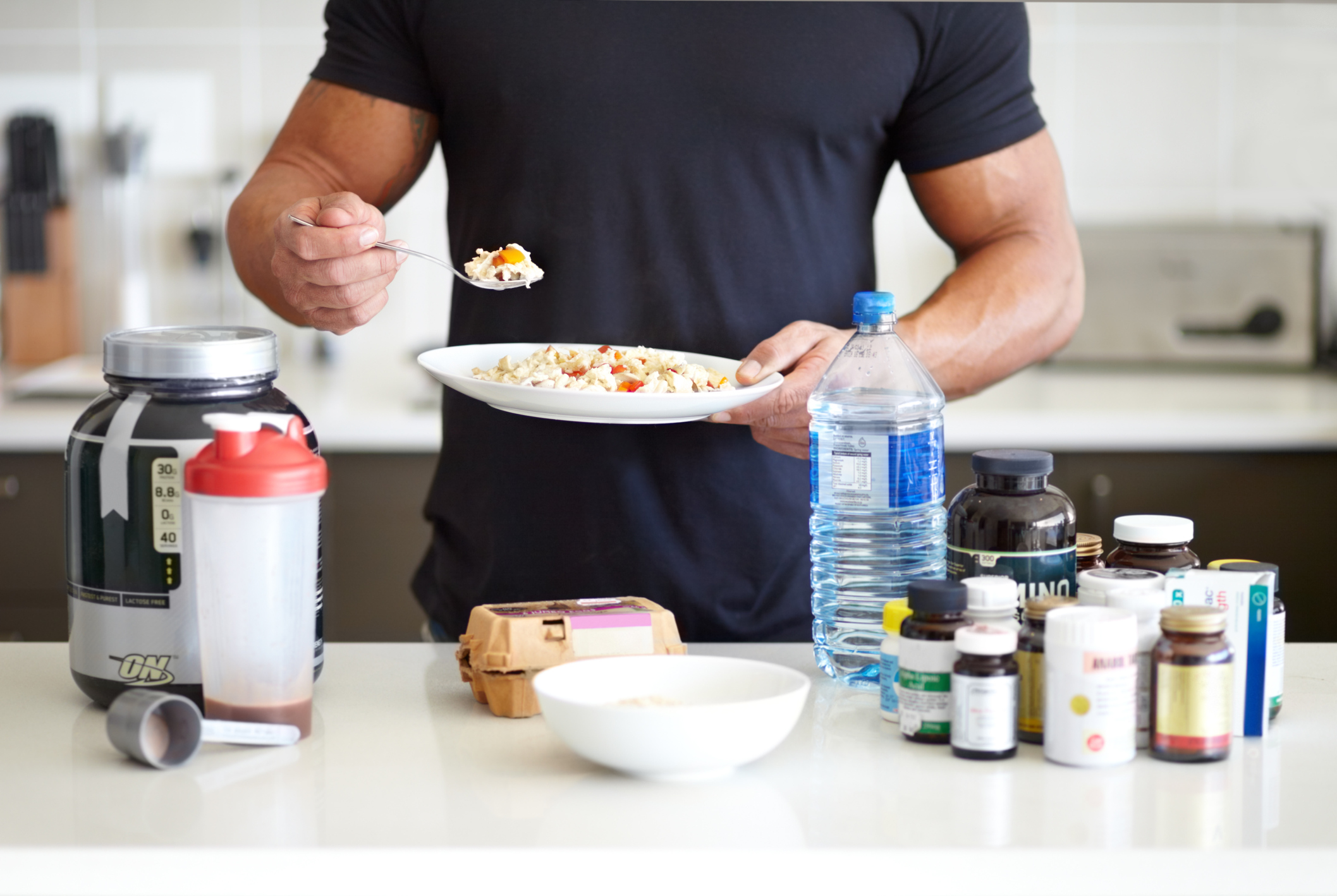 A muscular man holding a bowl of omelet while standing in front of his supplements