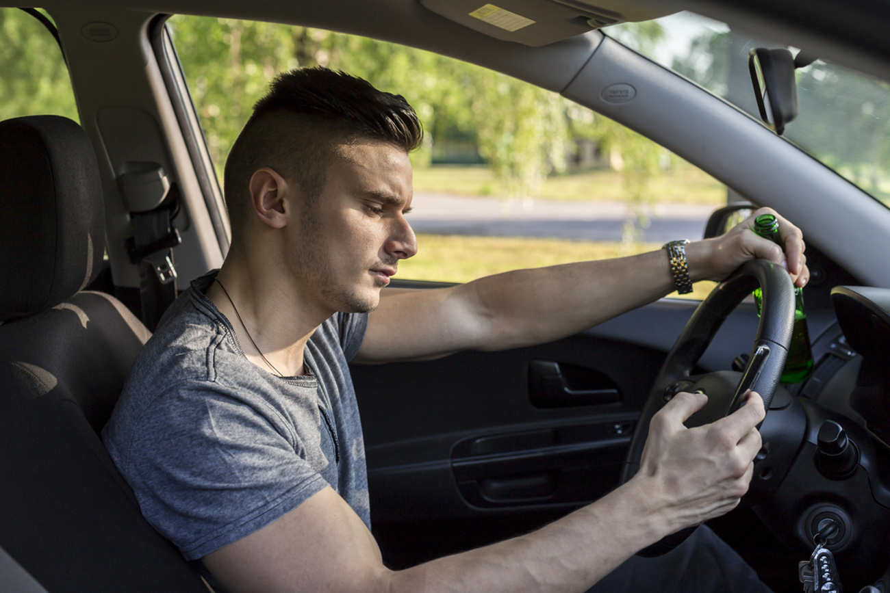 Young man drinking beer and looking at mobile phone while driving a car